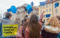 Młodzież stoi na rynku miasta i trzyma w rękach flagi europejskie.